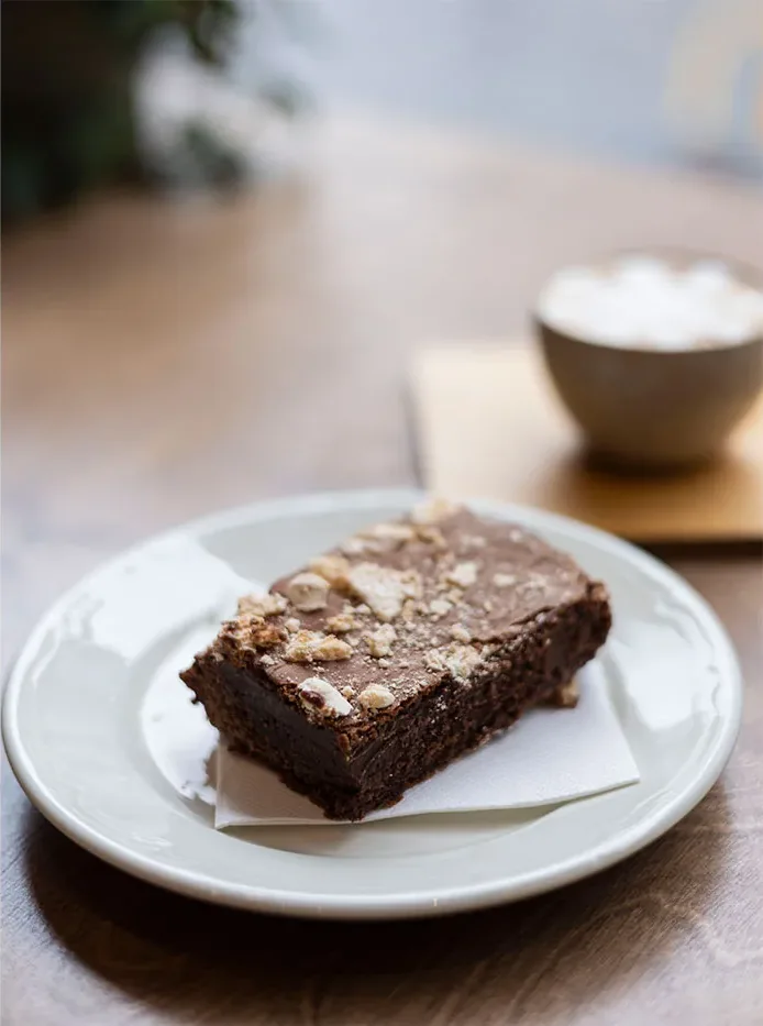 Une part de brownie au chocolat sur une assiette blanche, à côté d'un cappuccino