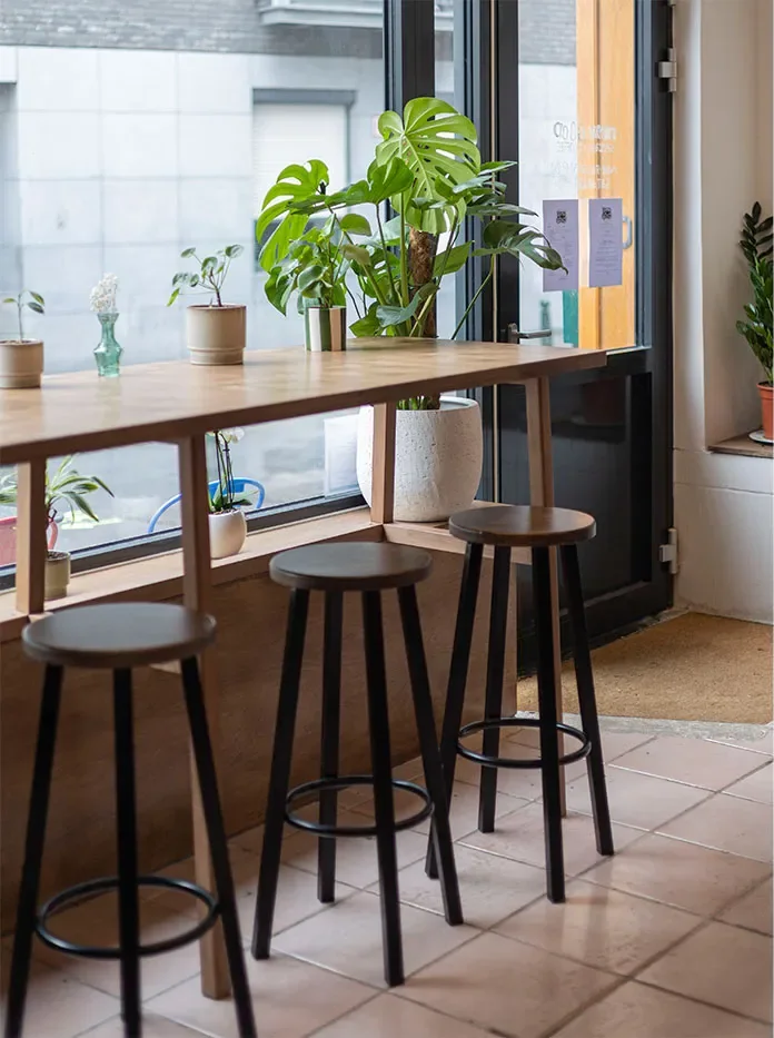 Trois tabourets hauts devant la tablette en bois qui longe la vitrine, plantes vertes au-dessus