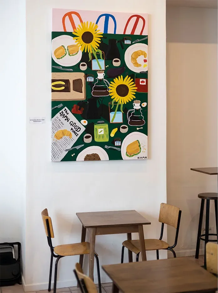 Une toile verte peinte de sandwichs, cafetières et tournesols, accrochée près de deux tables d'écolier